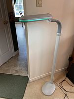 Side view of the white OttLite floor lamp showing the elongated light head with green accent strip, flexible neck, and oval base on carpet.