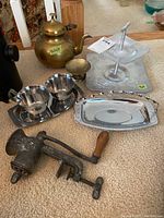 Photo showing vintage metal ware including brass teapot, hand-crank meat grinder, metal cream and sugar set with tray, egg cups, small cup, and two metal trays arranged on carpet.