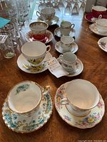 Photo showing eight assorted cups with saucers on wooden table, featuring floral patterns and gold rim details, some with visible wear and one chipped demitasse cup