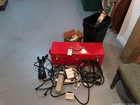 Photo showing red metal toolbox, black extension cords with power bars, and a black waste bin in basement floor area.