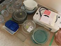 Wide view of multiple glass cookware pieces stacked and grouped, including clear Pyrex bowls with lids, blue covered dish, and white opaque mixing bowls with floral pattern.