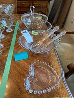 Seven pieces of vintage glassware on a wooden table including etched heart-shaped dish, cake plate with bead rim, large bowl with matching plate, two serving utensils, and cornflower salad bowl.
