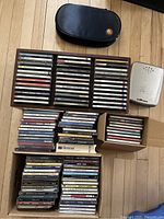 Photo showing the wooden CD storage box filled with CDs, two black travel CD cases, and a silver CD player or case on the side, all displayed on a wooden floor.