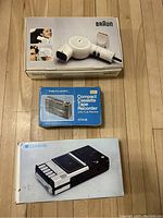 Three vintage electronics still in their original boxes: a Braun hair dryer, a Realistic compact cassette tape recorder, and a Lyons portable cassette tape recorder.
