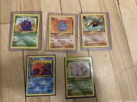 Five 1st Edition original Pokémon cards visible on wooden floor, each in a clear protective sleeve showing front art and details.