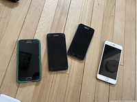 Four phones on wooden floor: two Samsung Galaxy S7 phones, one Samsung phone, and one rose-colored iPhone.