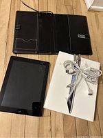 Photo showing iPad with cracked screen next to black folio case, original box, and charger cable.