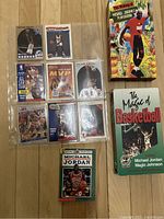 Photo shows 9 sleeved Michael Jordan basketball cards laid flat on a wooden floor alongside a collectors book and two VHS tapes.