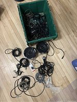Green plastic box containing several tangled microphone and audio cables on a wooden floor, with wired microphone and two speaker drivers outside the box.