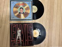 Top side of two Elvis Presley vinyl records with album covers: '3 Volume 3 Elvis Gold Records' and the 'Elvis Original Soundtrack from His NBC-TV Special'. Both records are partially removed from their covers and placed flat on a wooden floor.