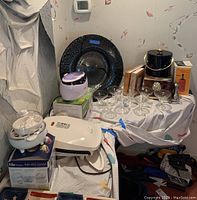 Full lot contents showing white George Foreman grill, ultrasonic jewelry cleaner, egg cooker in box, black decorative plate on stand, margarita glasses, metallic ice bucket, cookbooks and barware items.