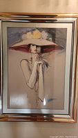 Full view of the framed artwork showing the woman wearing a large hat with flowers and a metallic frame.