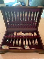 Open wooden box with top row of 12 knives and bottom row of assorted forks and spoons on maroon felt