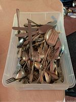 Top-down photo of clear plastic container full of mixed flatware including forks, spoons, knives, and serving pieces with various handles and designs, placed on a pink tablecloth.