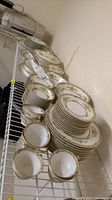 Wide view of stacked and arranged Noritake Alicia dinnerware showing multiple plates, bowls, and cups on wire shelving.