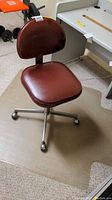 Brown leather-like office chair positioned on transparent plastic floor mat on carpeted floor.