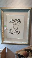 Full view of the framed abstract wall art featuring a minimalist face in black ink on a light background, with textured light frame.