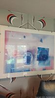 Wall art mounted on board with acrylic clipped front, showing geometric blue shapes blending with pink hues in an abstract style.