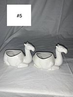 Two white camel-shaped sugar cube bowls placed side by side on a white cloth background, showing their profile and front orientation.