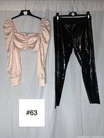 Photo of both baby pink satin long sleeve cropped top and black vinyl leggings hanging on black hangers against a white background with lot number sign.