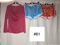 Pink long sleeve cowl neck shirt and two pairs of shorts (blue and pink/orange Hawaiian print) hung on hangers against a white background showing the full items.