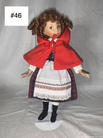 Full view of the porcelain doll dressed as Little Red Riding Hood, standing on white stand against grey background.