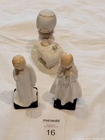 Three figurines on white background: two Royal Doulton figures in white gowns on black bases (Bedtime, Darling) and one Precious Moments figure of a child leaning on a bed