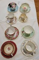 Top-down view of all 8 tea cups with their saucers arranged on a white cloth, showing various floral and gold patterned designs.