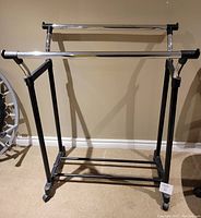Photo of metal adjustable rolling clothes rack with two horizontal chrome bars and black metal legs. Rack features wheels for mobility and adjustable height mechanism.