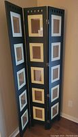 Full view of the tri-fold black wooden picture frame standing against a wall, showing the 15 photo openings with mats.