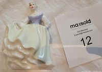 Porcelain figurine of a lady in a flowing gown, posed with one arm extended and wearing a purple bodice. MaxSold lot tag visible next to figurine.