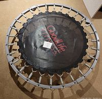 Top view showing the round fitness trampoline's metal springs around the perimeter connecting the black jumping mat to the frame, missing spring cover, and torn rubber on one leg.