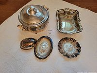 Four silver plated serving pieces including footed dish, wine tray with cut glass insert, serving dish with enamel pot, and butter dish with cut glass insert, arranged on white cloth.