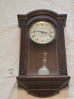 Full view of the wooden battery operated wall clock lying flat showing the clock face with Roman numerals, pendulum behind glass, and carved decorative features on the frame.