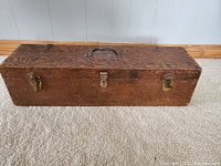 Full view of rectangular oak carpenter's toolbox on floor with three metal latches and metal handle on top.