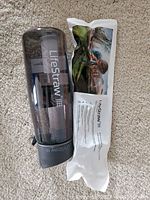 Photo showing the clear plastic Lifestraw water bottle with grey lid and visible filter inside. Next to the bottle is a sealed filter cartridge in its packaging with outdoor nature imagery and Lifestraw branding.