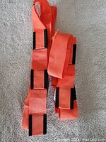 Two orange forearm forklift straps laid out on carpet, showing length and black reinforcement patches. Minor fraying visible on one strap's edge.