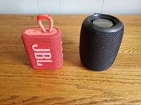 Photo showing red JBL GO3 Bluetooth speaker and black Creative Bluetooth speaker side by side on wooden surface.