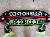 Photo showing both scarves laid flat on carpet, the top black scarf with COACHELLA text in white and red stripe border, bottom scarf green and black with GLASGOW CELTIC text, shamrocks and "THE PRIDE OF PARKHEAD" text