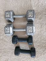 Four dumbbells arranged on carpet: two metal 25 lb hex dumbbells on top and two black vinyl 10 lb dumbbells below.