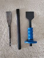 Three assorted chisels laid on carpet: Bosch SDS-plus chisel, flat metal hand chisel, and heavy-duty chisel with blue plastic handle.