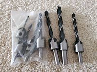 Three black twist drill bits with integrated countersink attachments and a plastic bag with extra drill bits in the background on carpet.