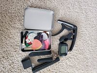 Lot items arranged on a carpet including a metal tin with colorful guitar picks, black capo, string winder, string trimmer, and digital tuner