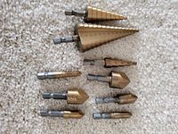 Photo showing an assortment of step cone drill bits and assorted bits arranged on carpet, highlighting two larger step cone bits at the top.
