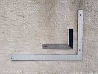 Large metal framing square and smaller metal carpenter square with black ABS set square, laid out on beige carpet.