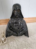 Front view of glossy black ceramic Darth Vader night light standing on carpet, showing helmet, mask, and illuminated chest piece area with power cord visible.
