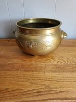 Photo showing the rounded brass bowl with dragon etched relief on the side and animal head handles, set on wooden surface with neutral background.