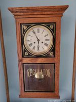 Front view of the oak regulator wall clock showing the clock face with Roman numerals and 'REGULATOR' text below on the glass panel with visible pendulum.