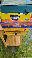 Stack of six board and party game boxes, titles visible including Feudal, Trivial Pursuit Party, Brainetics, The Worst-Case Scenario, AdVersity.