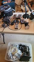 Photo showing table with multiple electronics and accessories including battery chargers, handheld device, power cables, two desk lamps, and carrying cases.